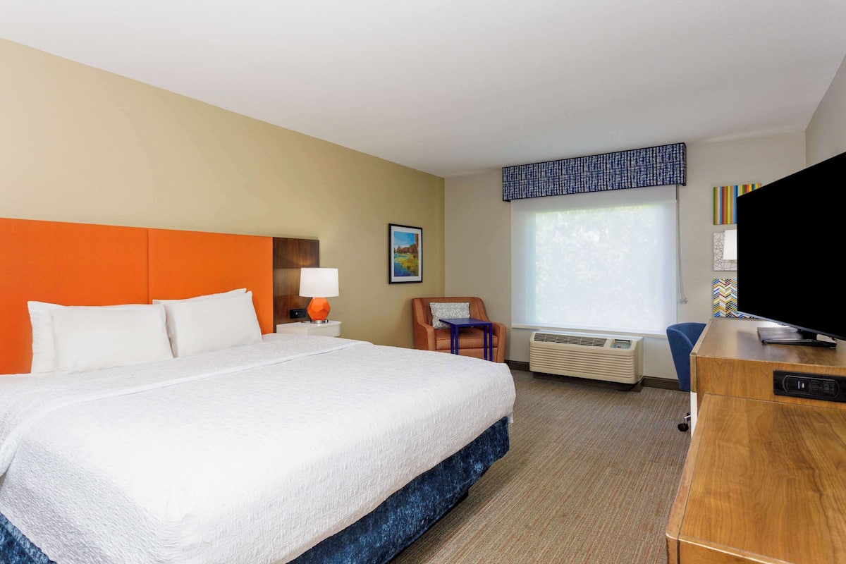 A hotel room with a large bed, a desk with a television, a chair, and a window with blinds.