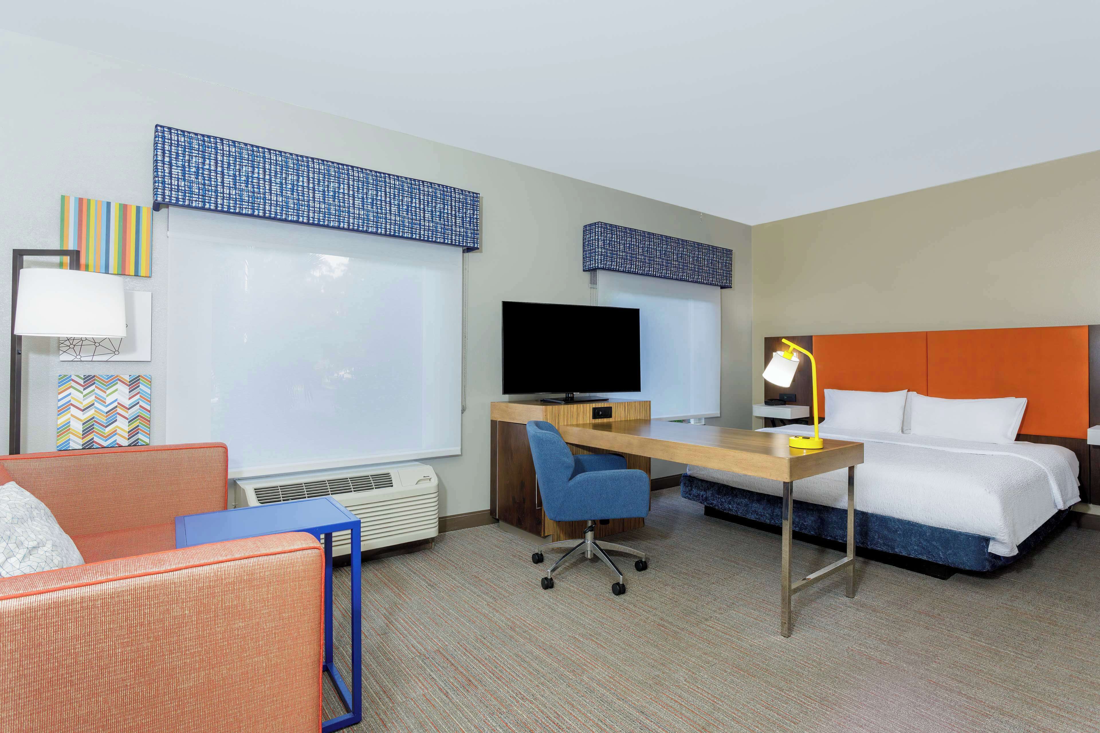 Une chambre d’hôtel comprenant un lit, un bureau avec un ordinateur, une chaise, une télévision et une fenêtre avec des stores.