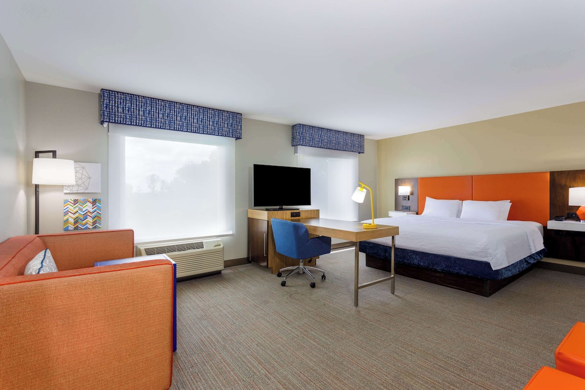 A hotel room with a bed, desk, chair, television, and a window with blinds.