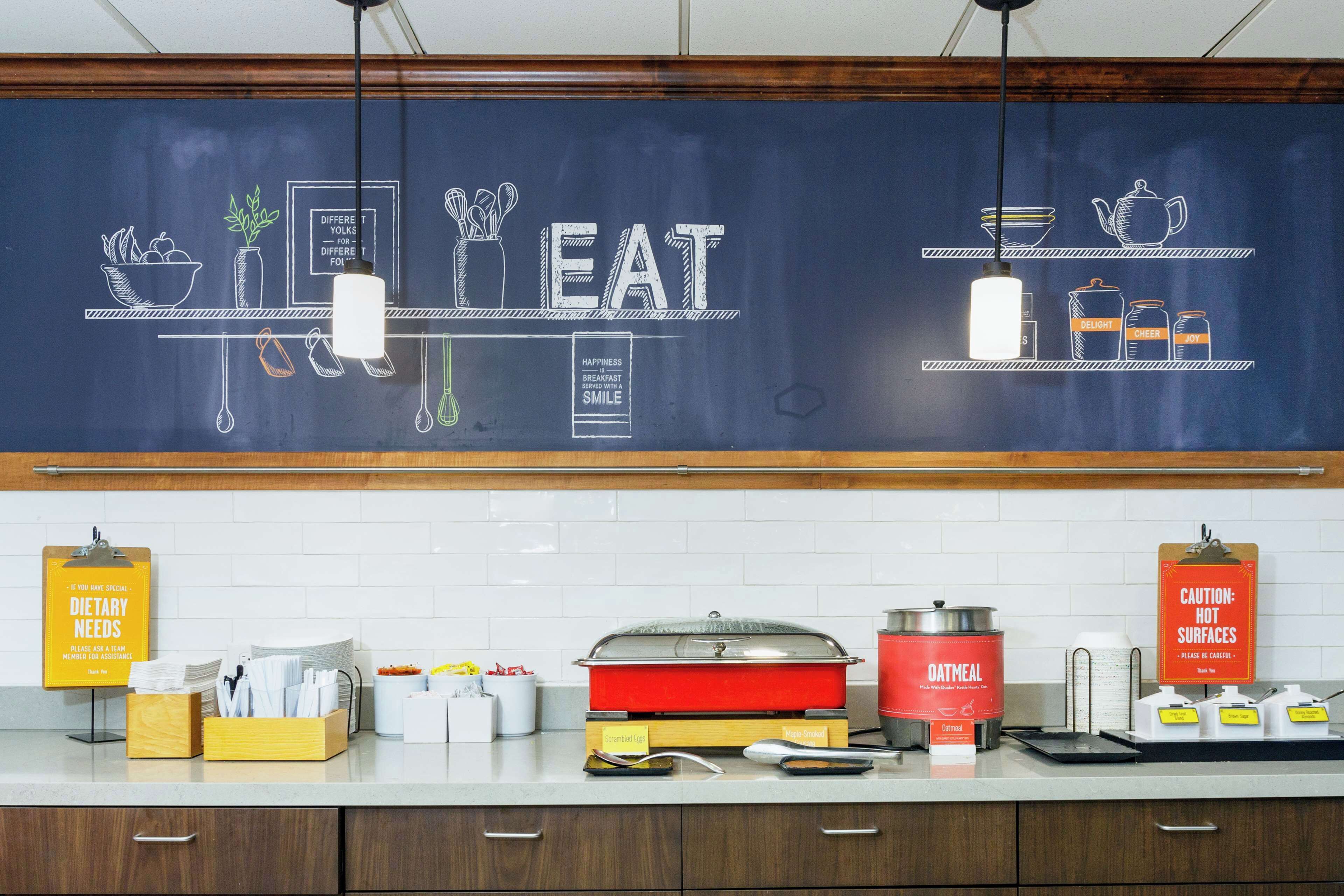 A kitchen with a chalkboard displaying food-related drawings and text, a red pot labeled "OATMEAL," and a sign indicating "CAUTION: HOT SURFACES."