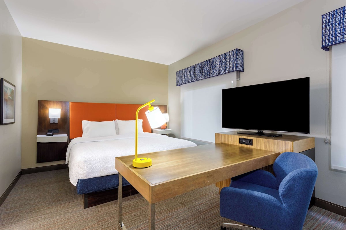 A hotel room with a bed, a desk, a chair, a TV, and a nightstand with a lamp.