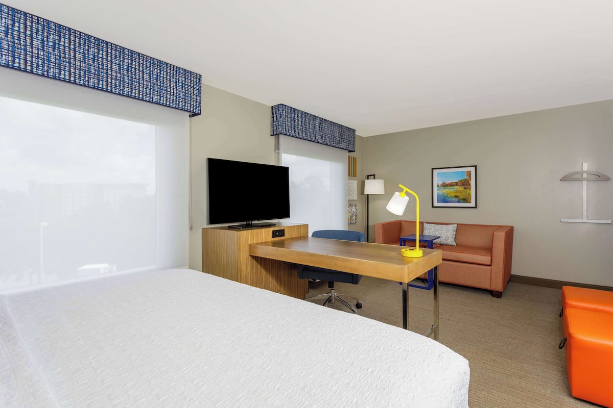A hotel room with a bed, a desk with a computer, a television, a sofa, and a small orange ottoman.
