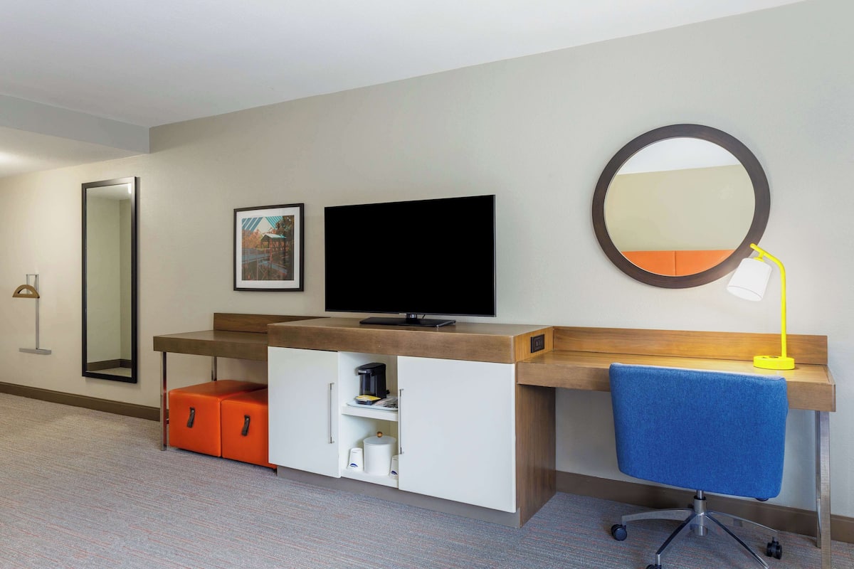 A hotel room with a flat-screen TV, a mirror, a desk with a chair, and a lamp.