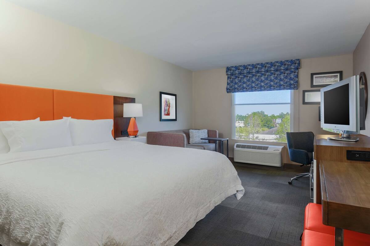 A hotel room with a large bed, a desk with a computer, a chair, a lamp, a window with a view, and an air conditioning unit.
