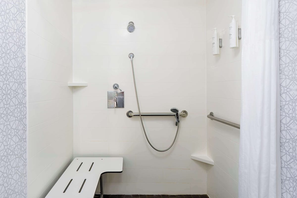 A shower area with white tiled walls, a handheld showerhead, a grab bar, and a white bench.
