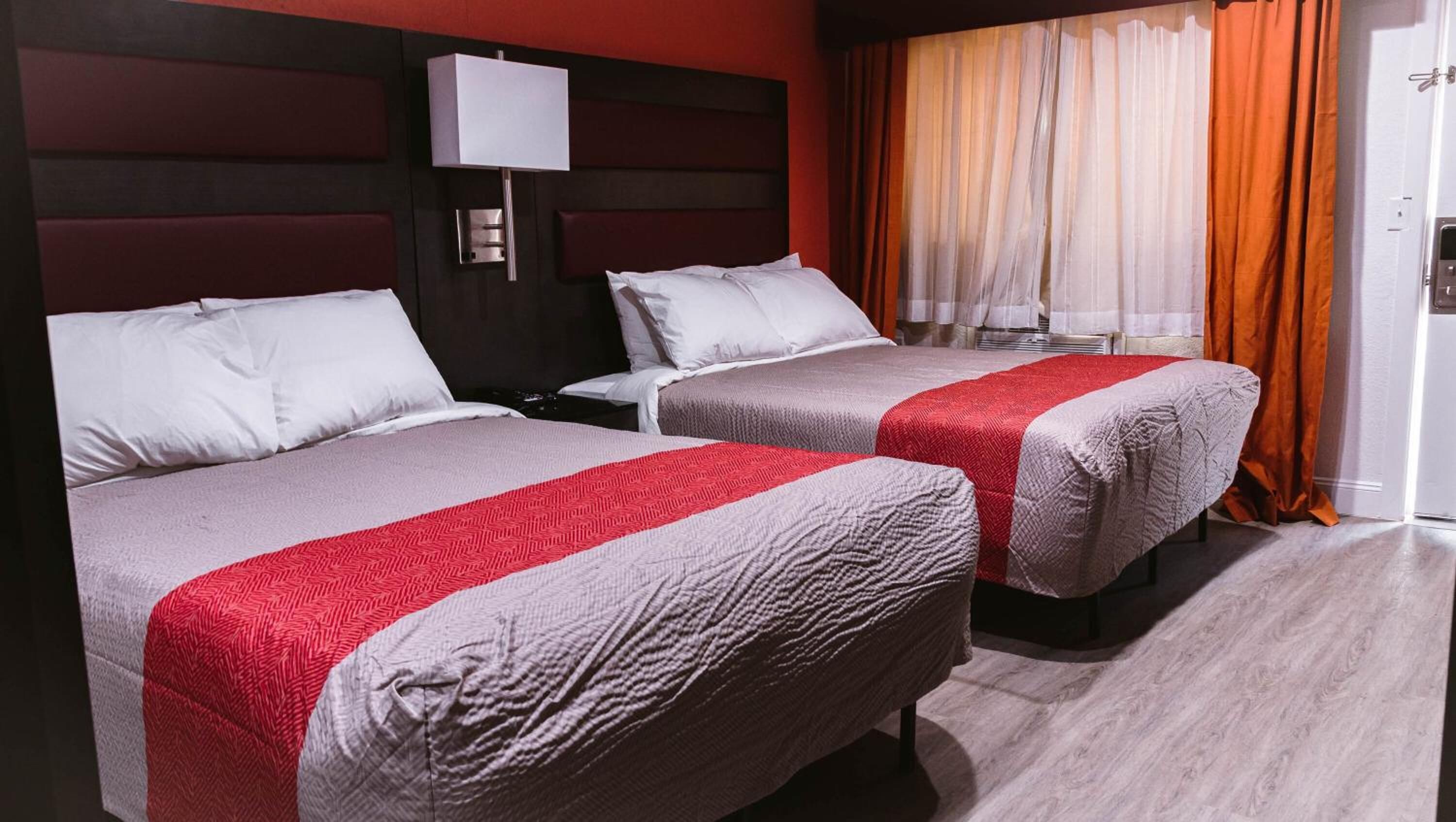 A hotel room with two beds, each with a red and grey bedspread, a nightstand, a wall-mounted lamp, and a window with orange curtains.