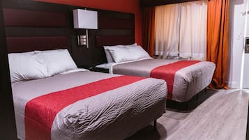 A hotel room with two beds, each with a red and grey bedspread, a nightstand, a wall-mounted lamp, and a window with orange curtains.