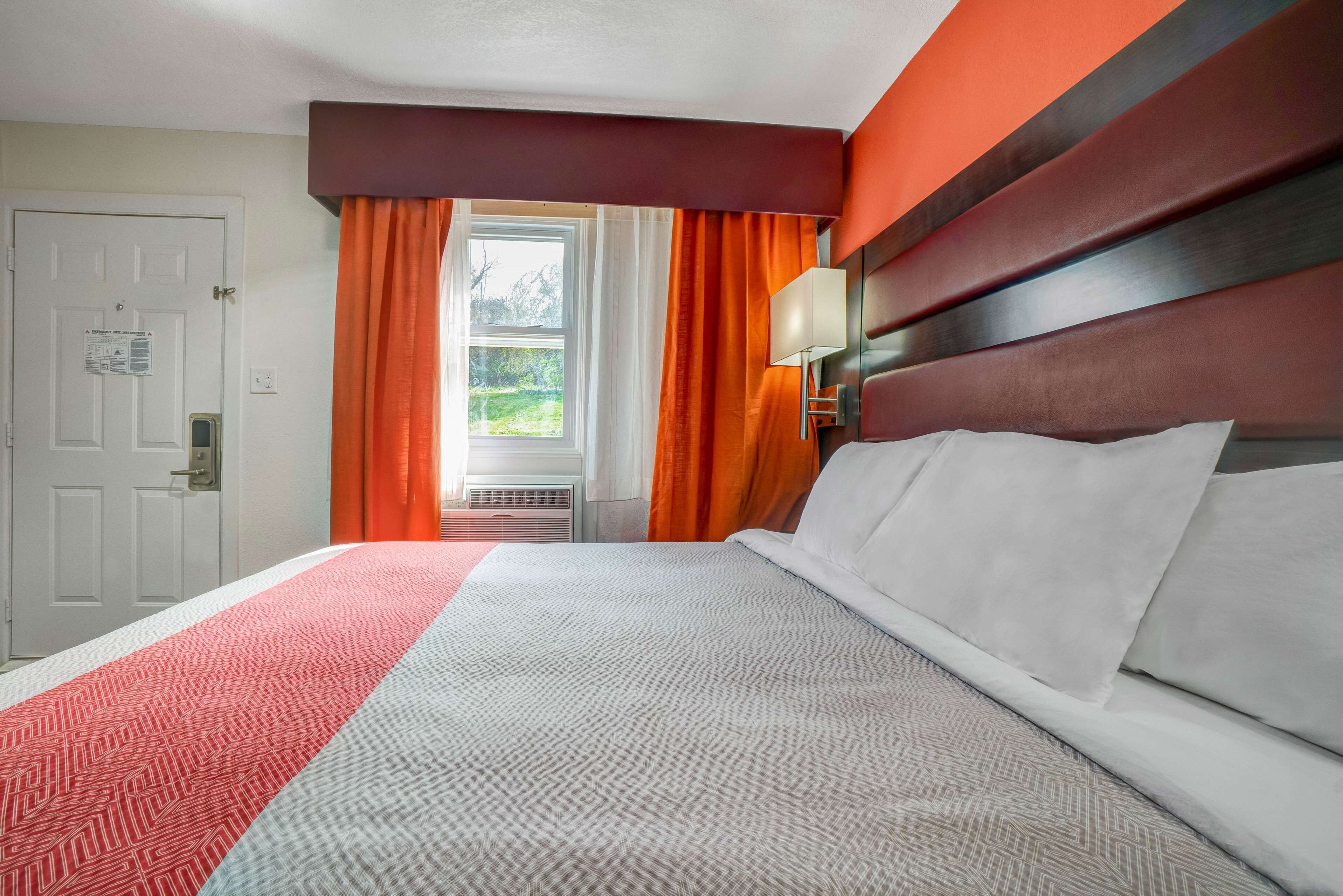 A hotel room with a bed, a window with curtains, a door, and a wall-mounted lamp.