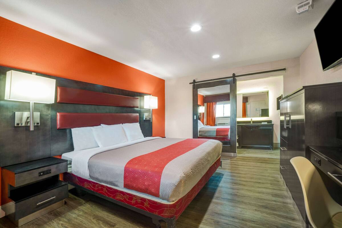 A hotel room with a large bed, bedside tables, a TV, and a bathroom visible through an open door.