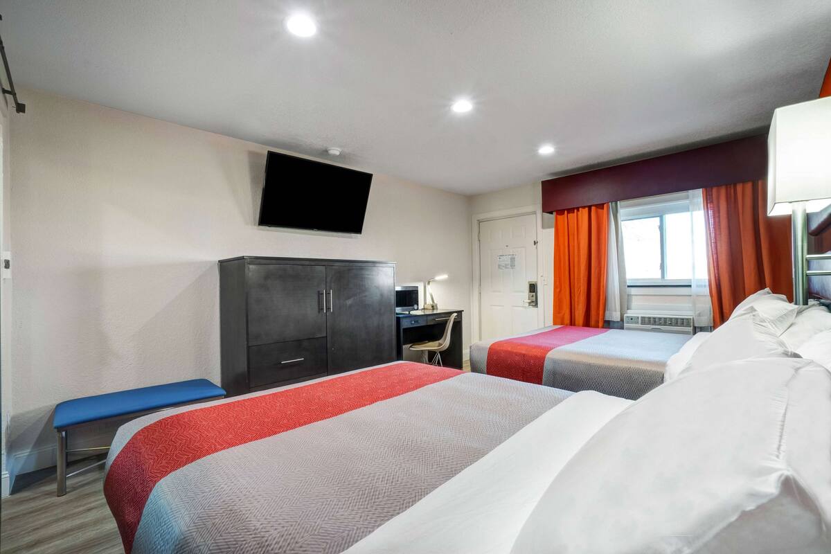 A hotel room with two beds, a desk, a TV mounted on the wall, and a window with orange curtains.