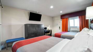 A hotel room with two beds, a desk, a TV mounted on the wall, and a window with orange curtains.