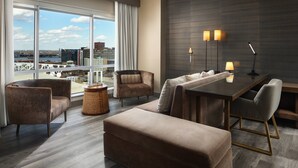 A modern hotel room with a large window offering a city view, a cozy seating area with a sofa and chairs, a dining table with lamps, and a wooden floor. - Louisville Marriott Downtown (Louisville)