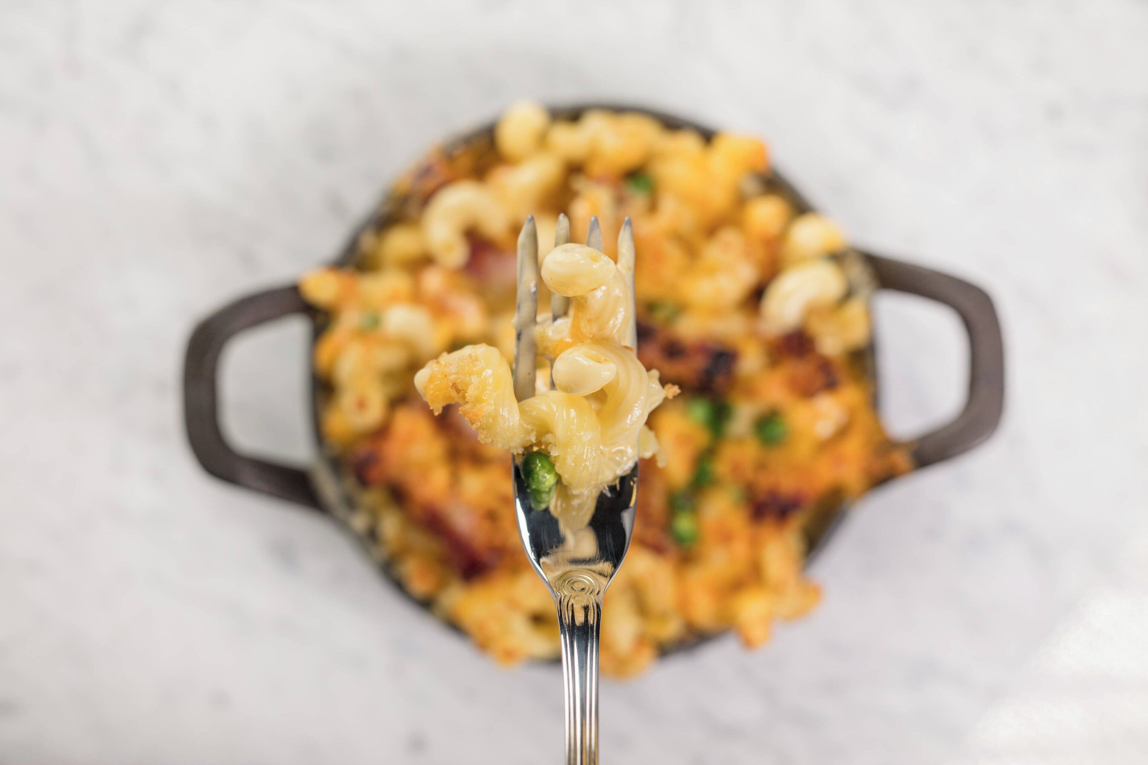 A close-up of a dish with macaroni and cheese, a fork lifting a portion, and a cast iron skillet in the background.