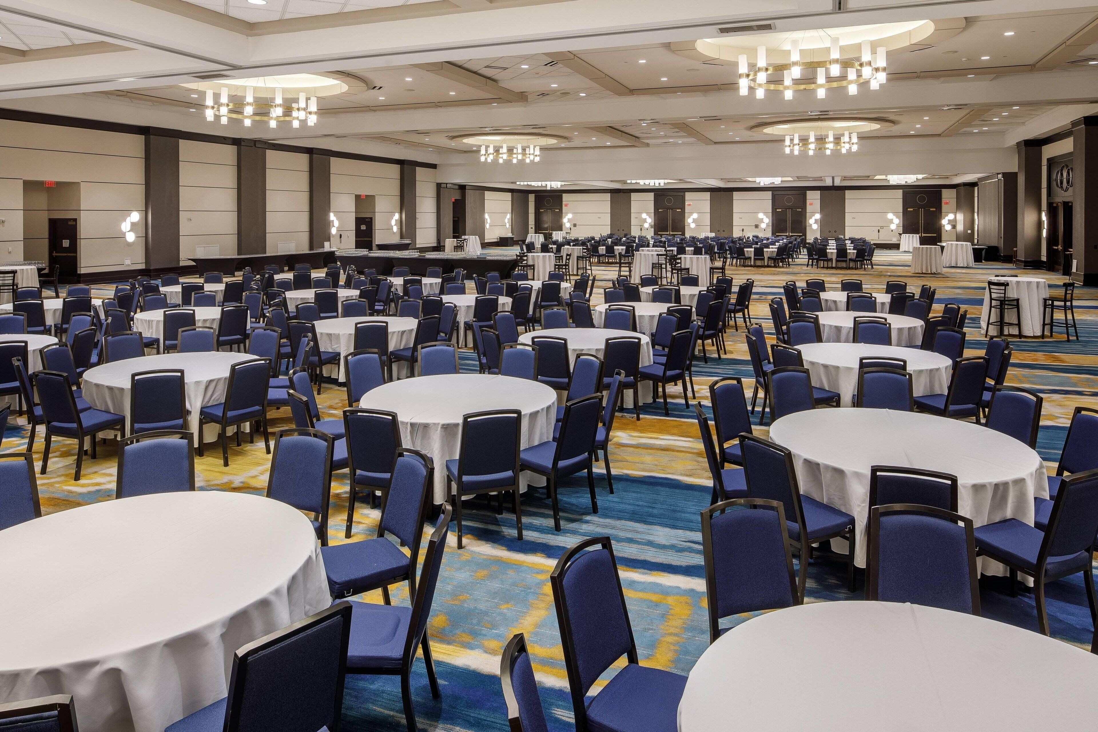 A large banquet hall with round tables and blue chairs, a stage, and a carpeted floor.