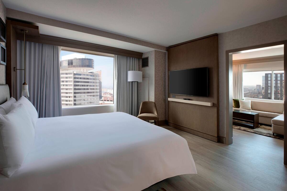 Ein Hotelzimmer mit einem großen Bett, einem flachbildfernseher an der Wand, einem Sessel, einer Couch und Blick auf die Stadt durch große Fenster.