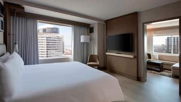 Habitación de hotel con una cama grande, un televisor de pantalla plana montado en la pared, una silla, un sofá y vistas de la ciudad a través de amplias ventanas.