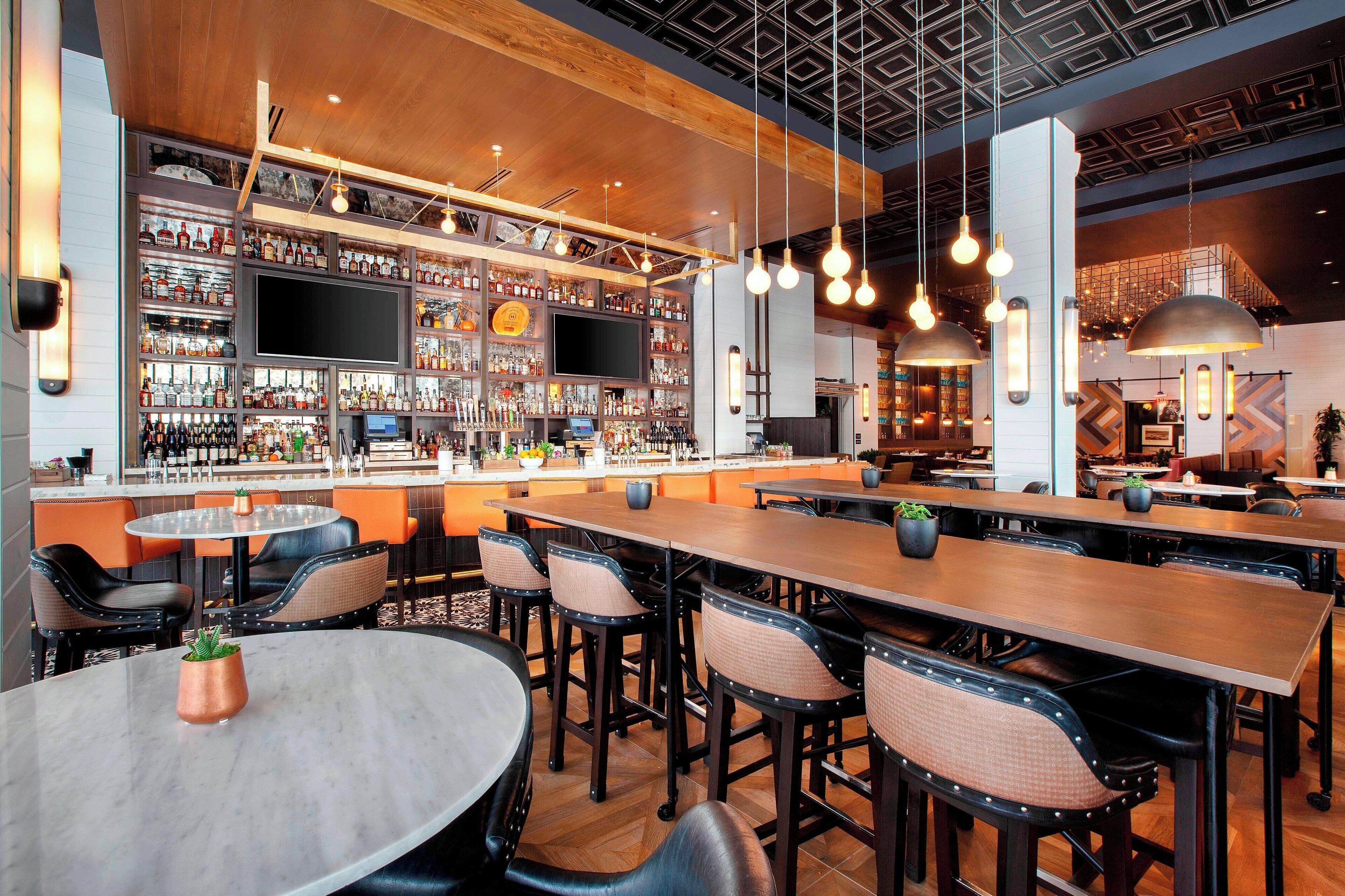 A bar area with multiple seating options, a television screen, and a variety of bottles on shelves.