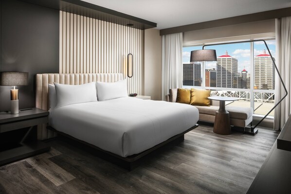 A modern hotel room with a large bed, a desk, a chair, a sofa, and a balcony with a city view.