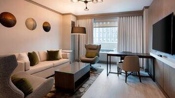 A modern hotel room with a sofa, armchair, coffee table, dining table, and a flat-screen TV.
