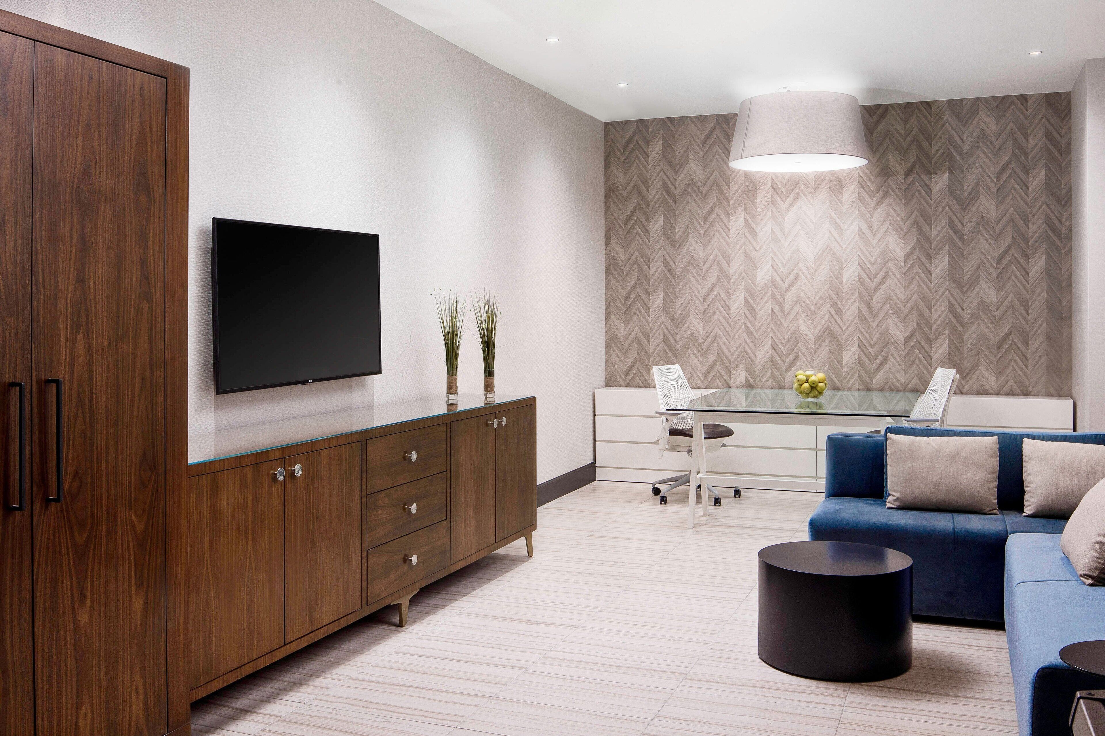 A modern living room with a large TV, a blue sofa, a glass coffee table, and a herringbone-patterned wall.