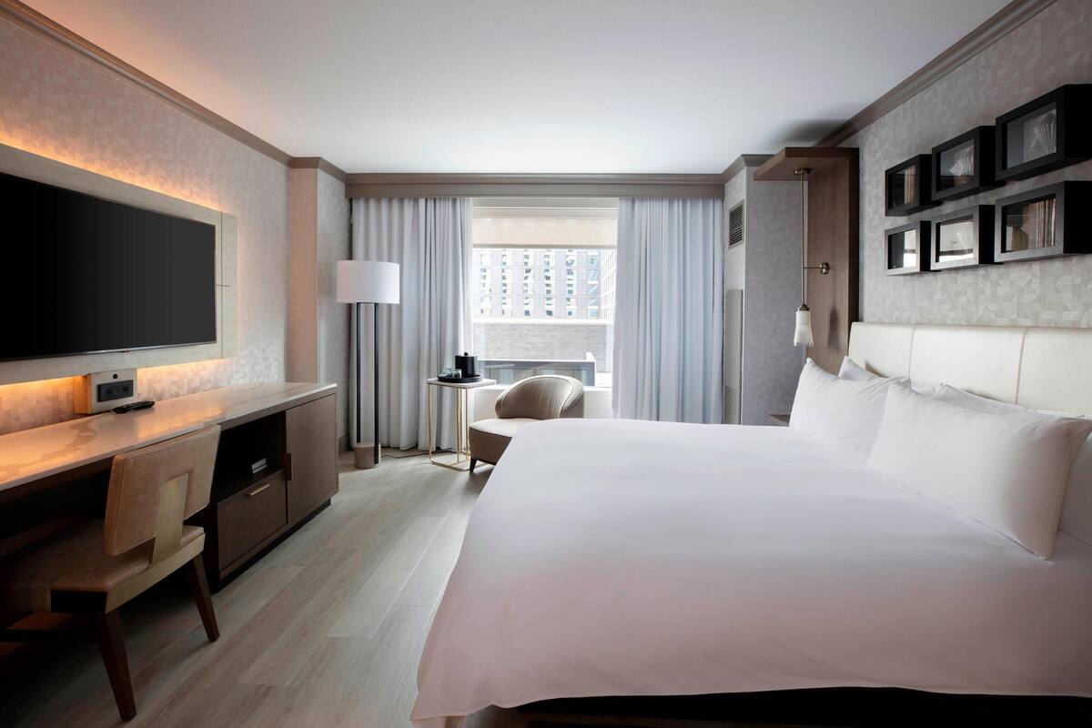 Ein Hotelzimmer mit einem großen Bett, einem Schreibtisch mit Fernseher, einem Sessel und einem Fenster mit Vorhängen.