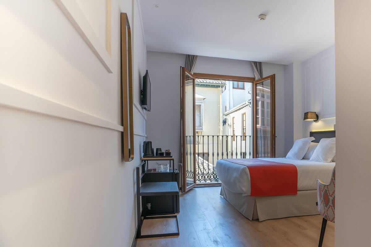 A hotel room with a bed, a desk, a chair, a TV, and a balcony with a railing.