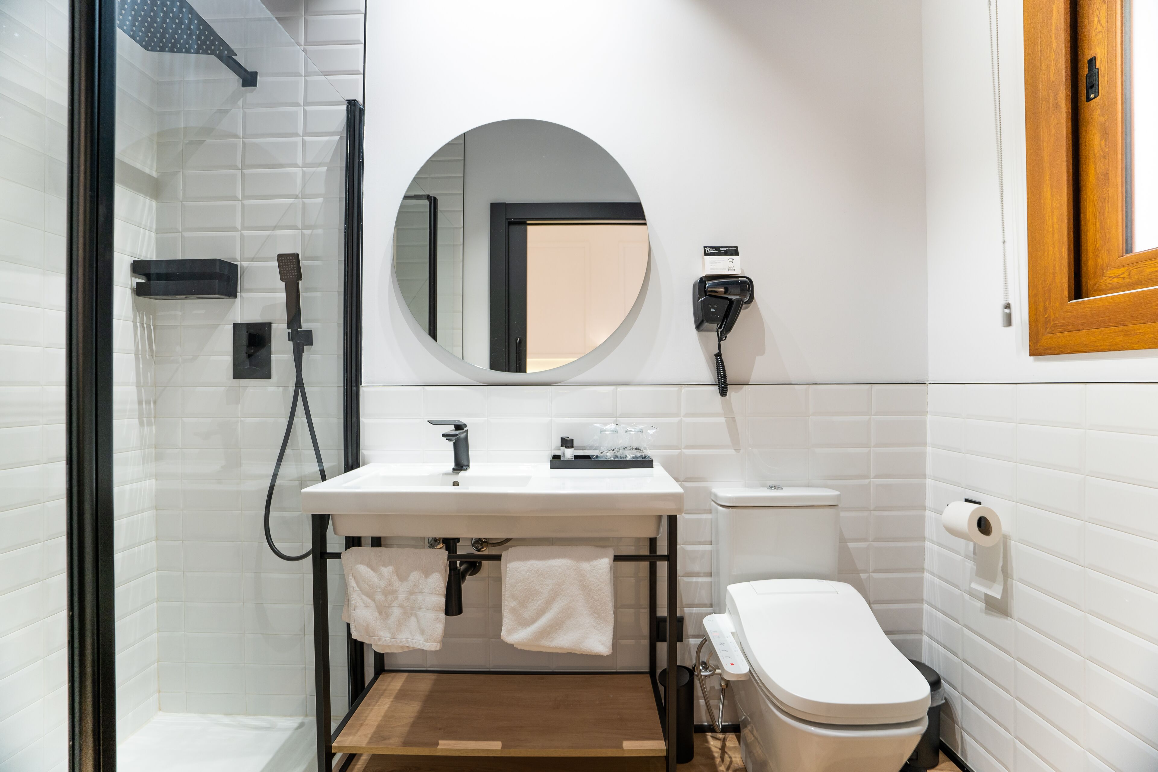 Un baño moderno con ducha de vidrio, un lavamanos blanco con espejo arriba y un inodoro con dispensador de papel higiénico.