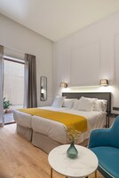 Una habitación de hotel con cama, mesita de noche, una silla azul, una pequeña mesa con un florero y vistas al exterior.