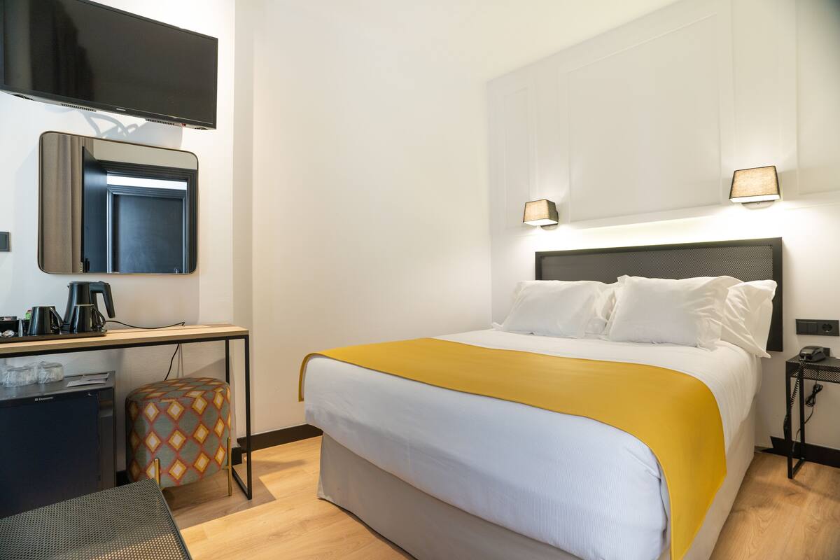 A hotel room with a bed, a desk, a chair, a small table, a television, and a nightstand with a lamp.