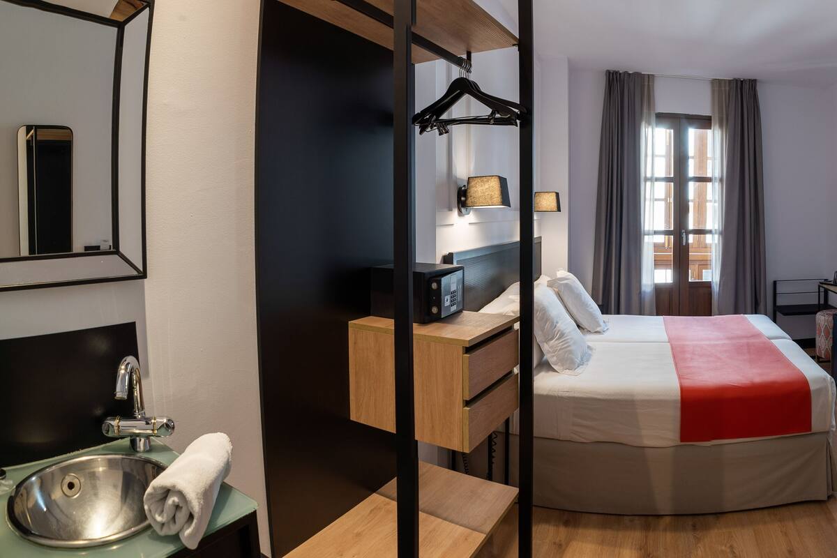 Habitación de hotel con cama, mesita de noche, espejo y vistas al exterior a través de una ventana.