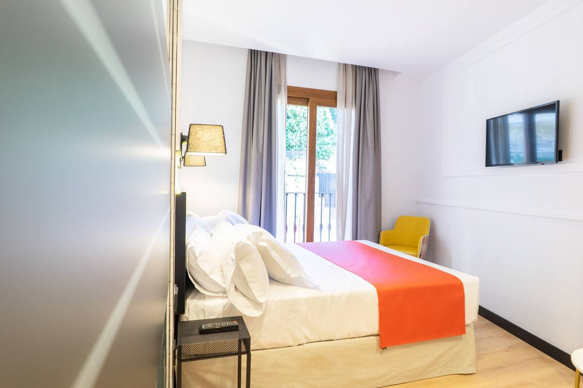 Una habitación de hotel con cama, una silla, televisor y ventana con cortinas.