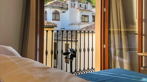 Habitación de hotel con balcón, cama, ventana con cortinas y vistas a edificios y un castillo a lo lejos.