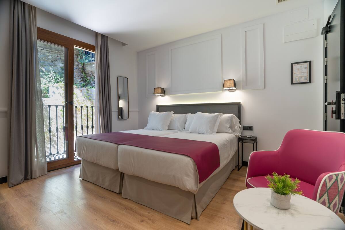 A double bed with a headboard, a small table with a plant, a pink chair, and a balcony with curtains.