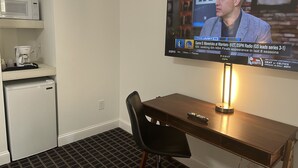 Ett hotellrum med en tv monterad på väggen som visar en sportmatch, ett litet kylskåp, en mikrovågsugn och en kaffebryggare på en hylla.