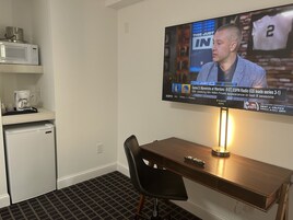 Habitación de hotel con una televisión en la pared mostrando un partido deportivo, un refrigerador pequeño, un microondas y una cafetera sobre una repisa.