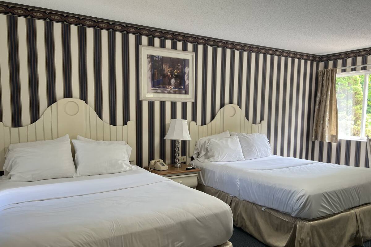 A hotel room with two beds, striped wallpaper, a painting on the wall, a bedside lamp, and a window with curtains.