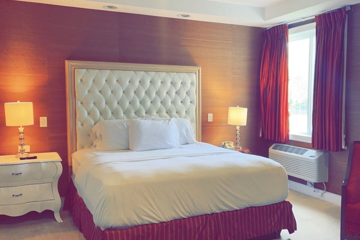 A hotel room with a large bed, two bedside tables, a red chair, a window with curtains, and a wall-mounted air conditioner.