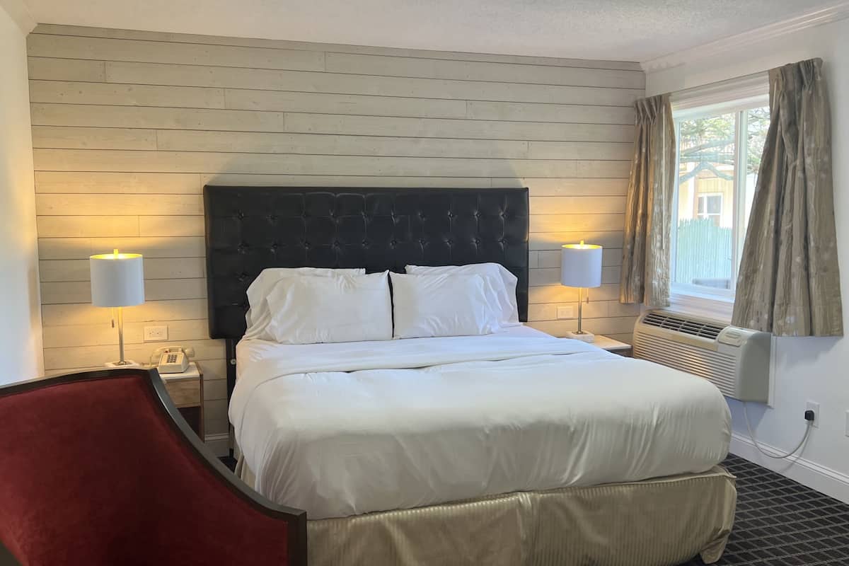A hotel room with a bed, two bedside lamps, a telephone, a red chair, and a window with curtains.