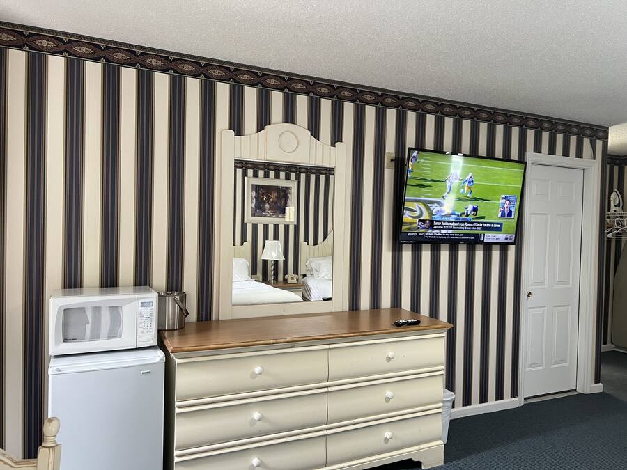 A hotel room with a striped wallpaper, a television mounted on the wall, a dresser with drawers, a microwave, and a door.