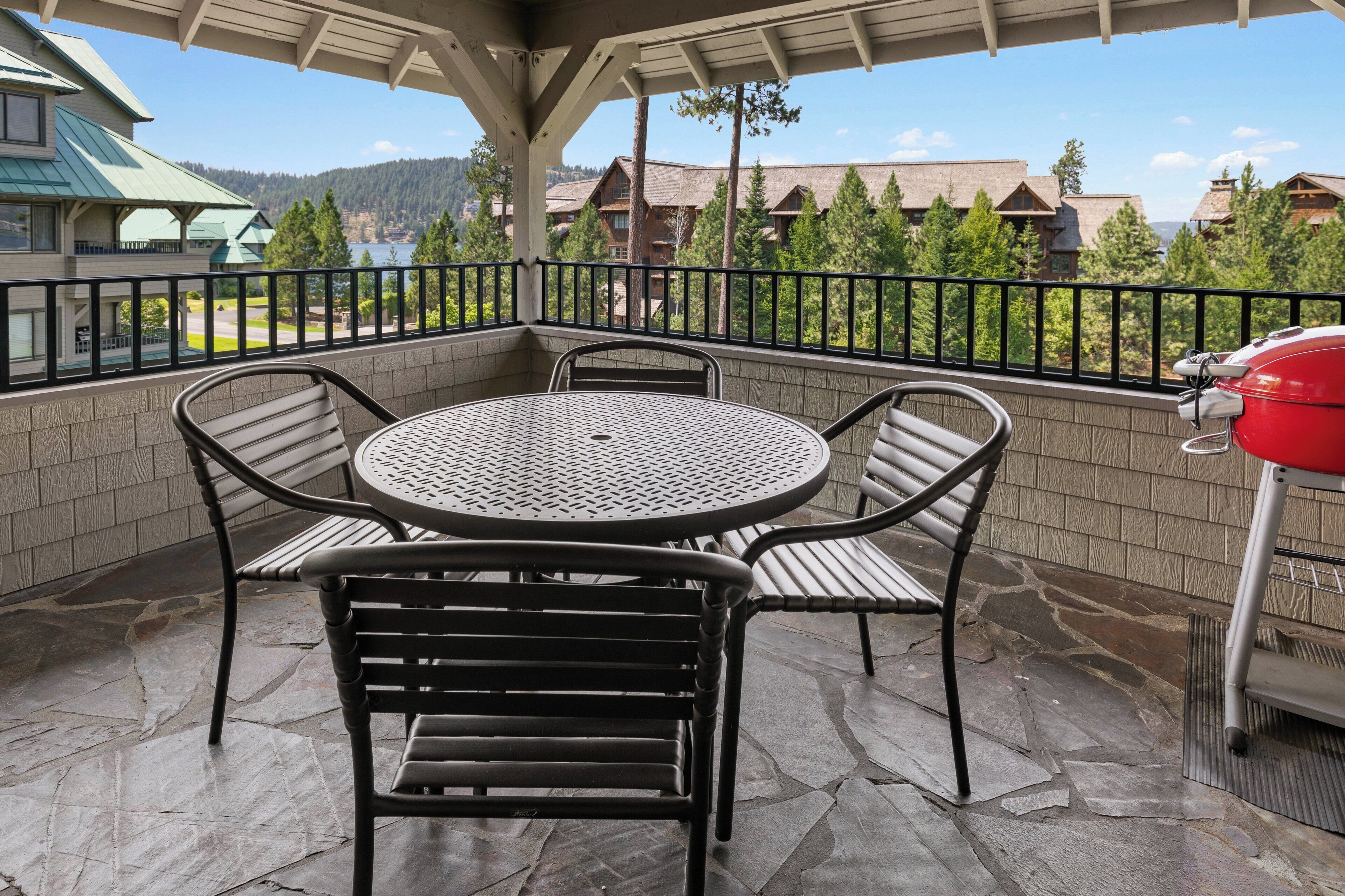 A covered patio with a round table and four chairs, a barbecue grill, and a view of buildings and trees in the background.