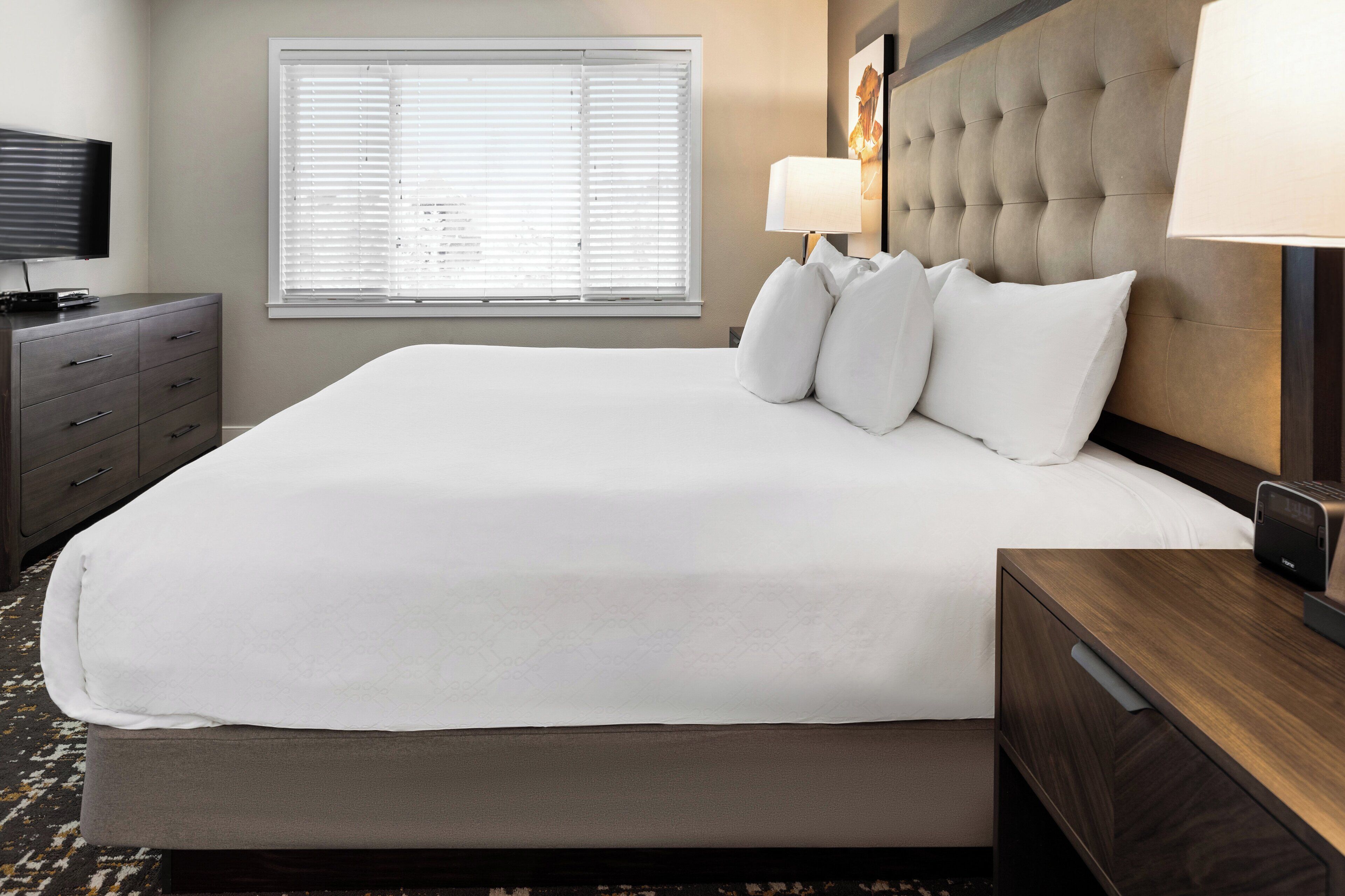 A neatly made bed with white linens, a tufted headboard, a bedside lamp, and a television on a dresser.