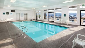 Una amplia piscina interior con paredes de vidrio transparente, sillas blancas y vistas a las montañas.
