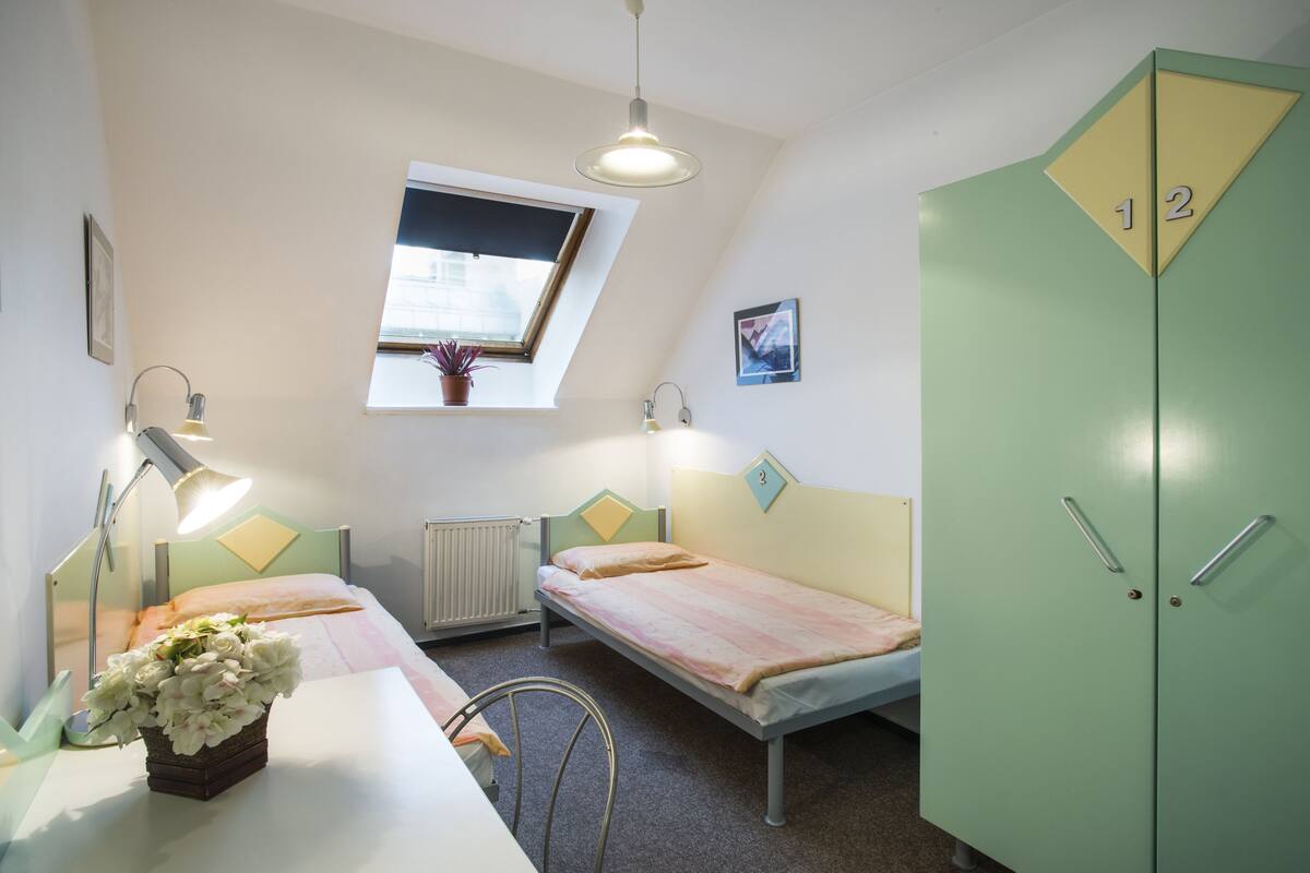 A dormitory room with two single beds, a desk with a lamp, a radiator, a window with a plant, and a wardrobe with numbered compartments.
