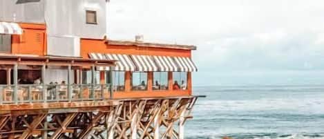 A coastal building with a deck extending over the water, featuring a balcony with seating.