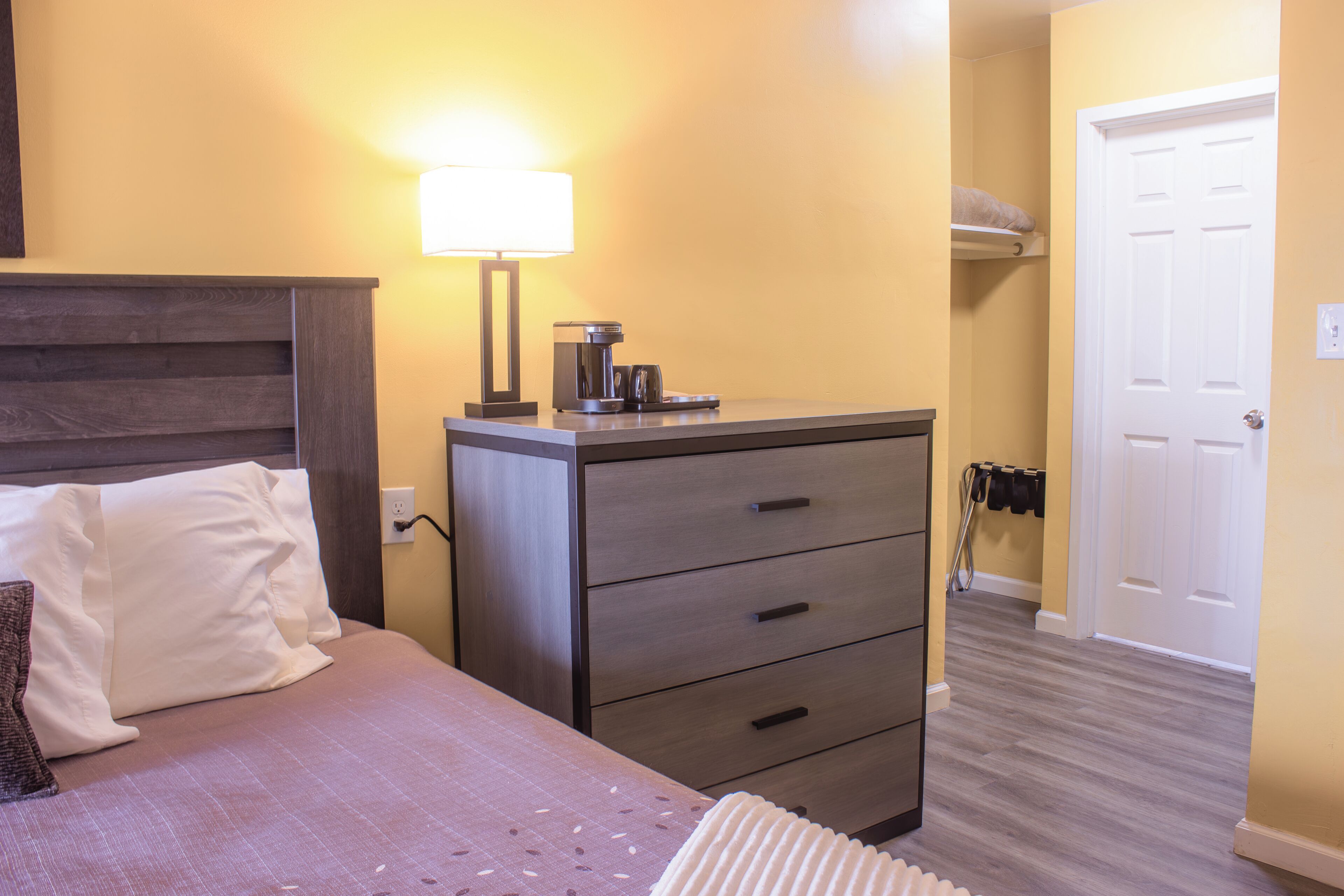 A hotel room with a bed, a nightstand, a lamp, a coffee maker, and a wardrobe.