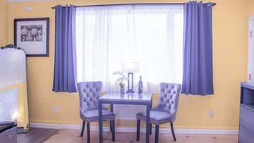 A dining area with a table and two chairs, a window with curtains, a framed picture on the wall, and a small table with a lamp and wine glasses.