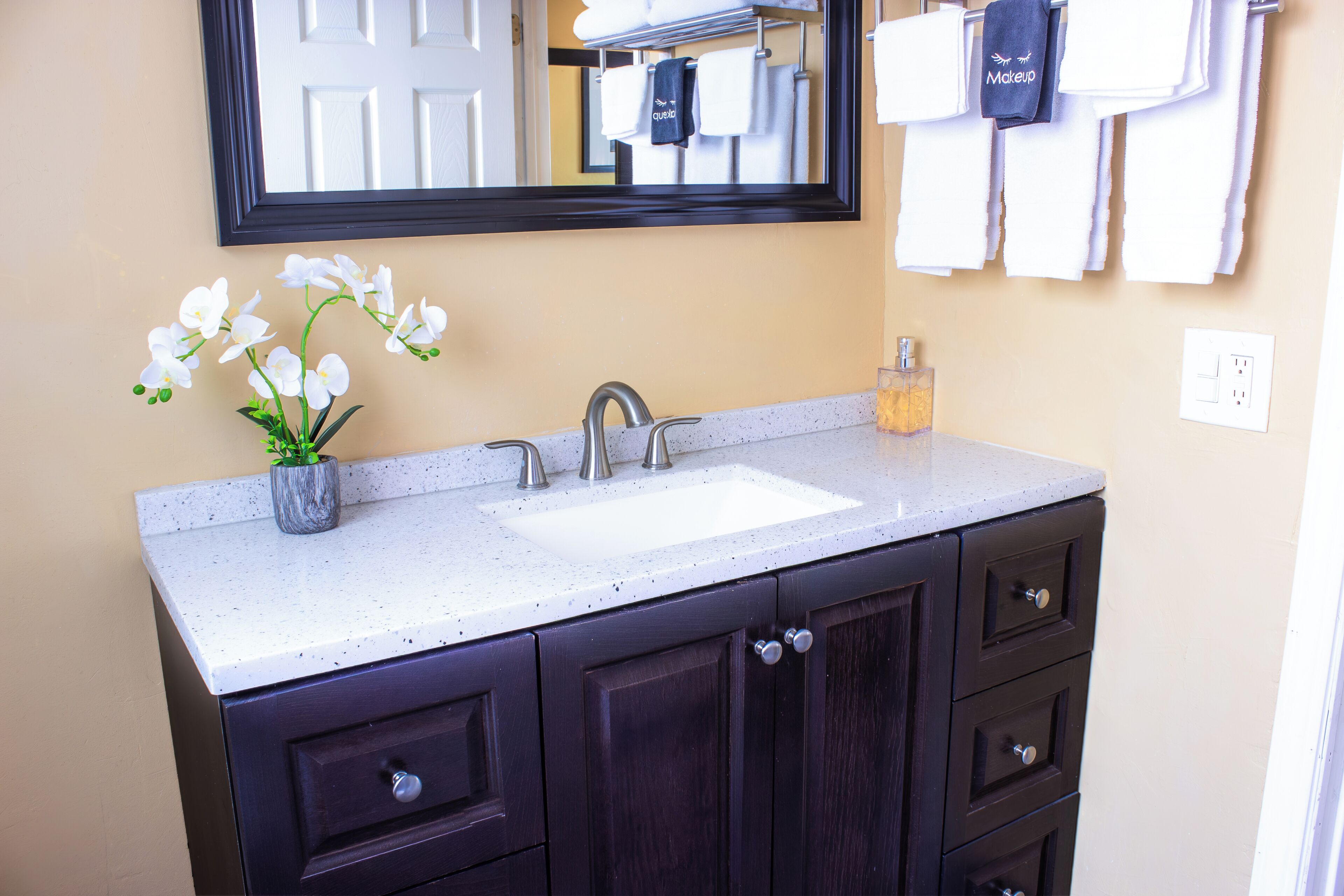 Un baño con lavamanos blanco, muebles de madera oscura, un espejo sobre el lavamanos y una orquídea en maceta en la encimera.