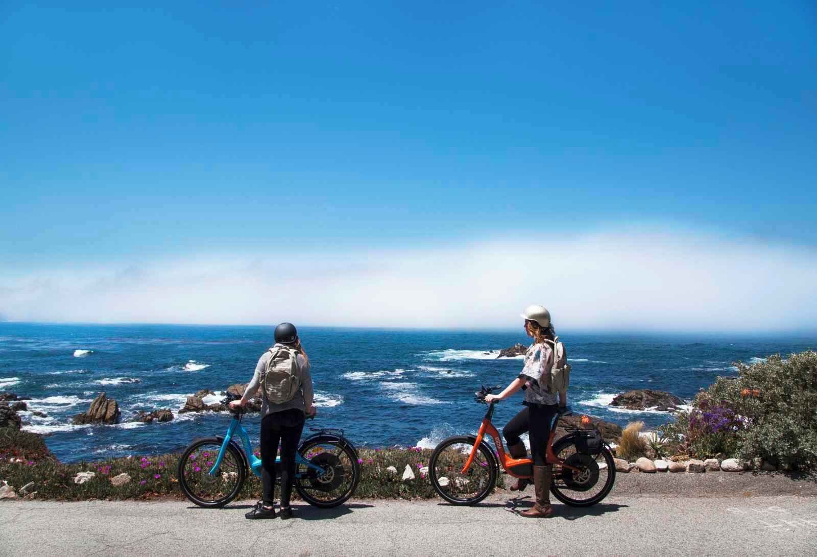 Dos personas en bicicleta por una carretera costera, con vistas al océano.