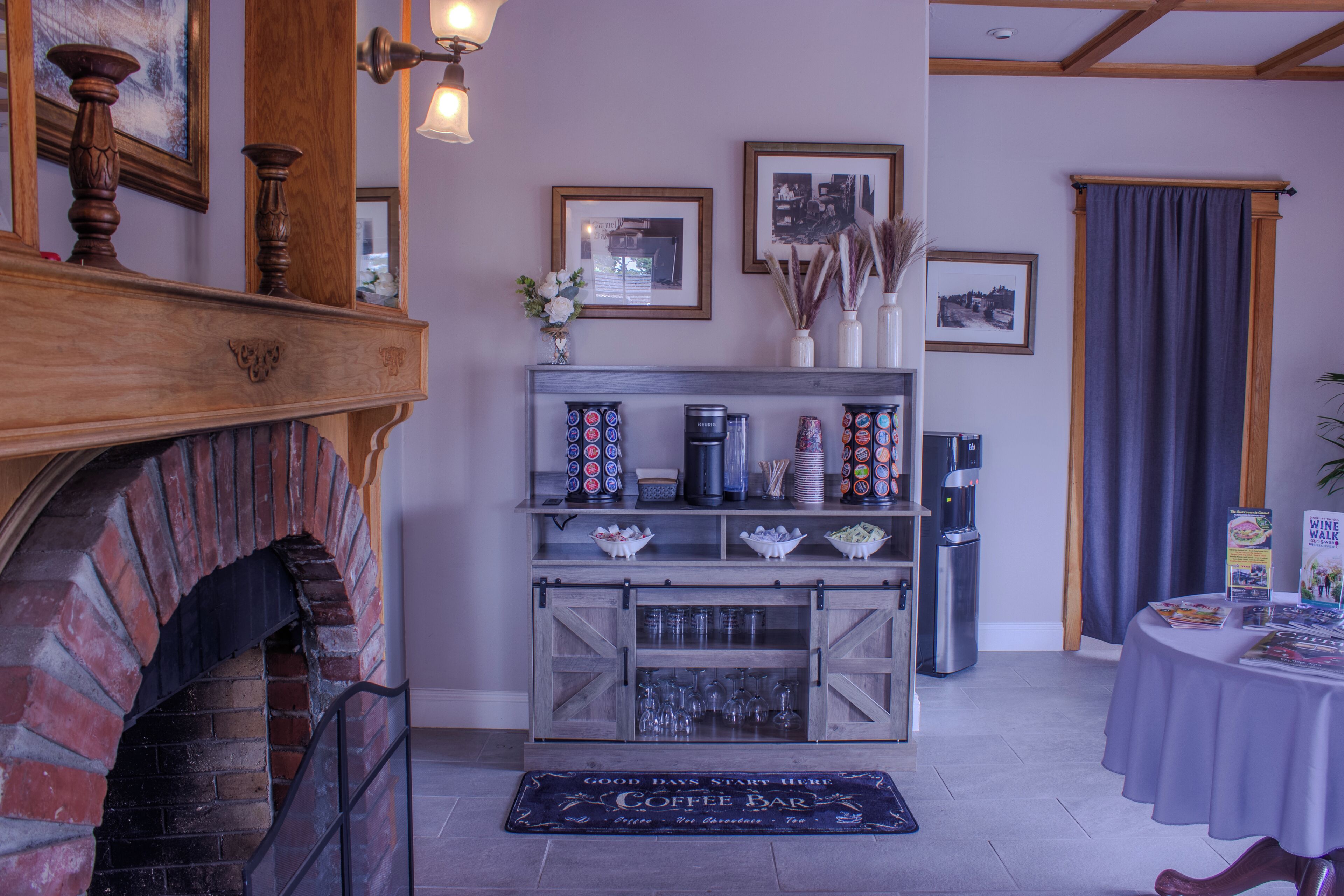 A cozy room with a brick fireplace, a coffee bar area with various mugs and a coffee machine, and a wall decorated with framed pictures and a clock.