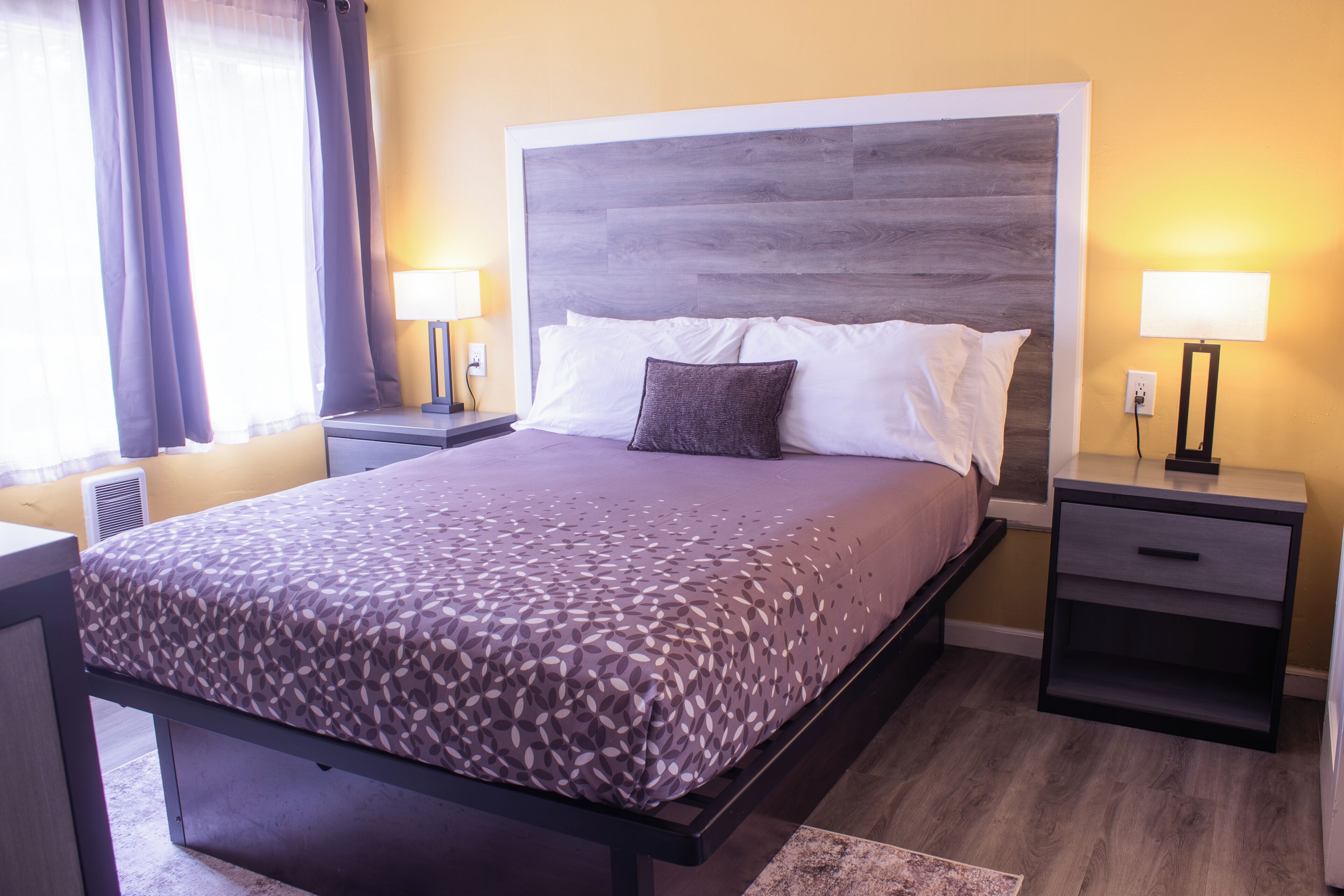 A hotel room with a large bed, two bedside tables, a nightstand, a wall-mounted lamp, and a window with curtains.